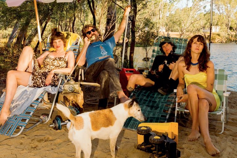 Australian rock band Tropical Fuck Storm lounging on lawn chairs outdoors by a riverbank with a dog. Photo taken by Jamie Wdziekonski.